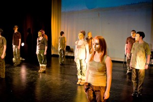 IMAGE Students stand on a lit stage space wearing theatrical gold masks IMAGE