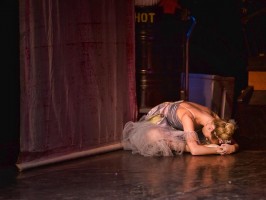 A dancer in mauve rests her head on the stage floor as she stretches forward over crossed legs.
