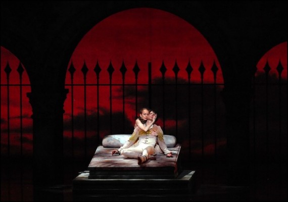 IMAGE Photo by Michael Seamans - Dancers: Larissa Ponomarenko and Nelson Madrigal (Boston Ballet) - Romeo and Juliet embrace on a bed before a red backdrop. IMAGE