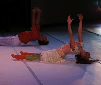 [image] Two dancers lying on the floor, lift their backs into an arch as they reach upward [image]