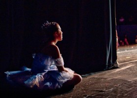 A young ballerina in purple tutu sits watching from the wings.
